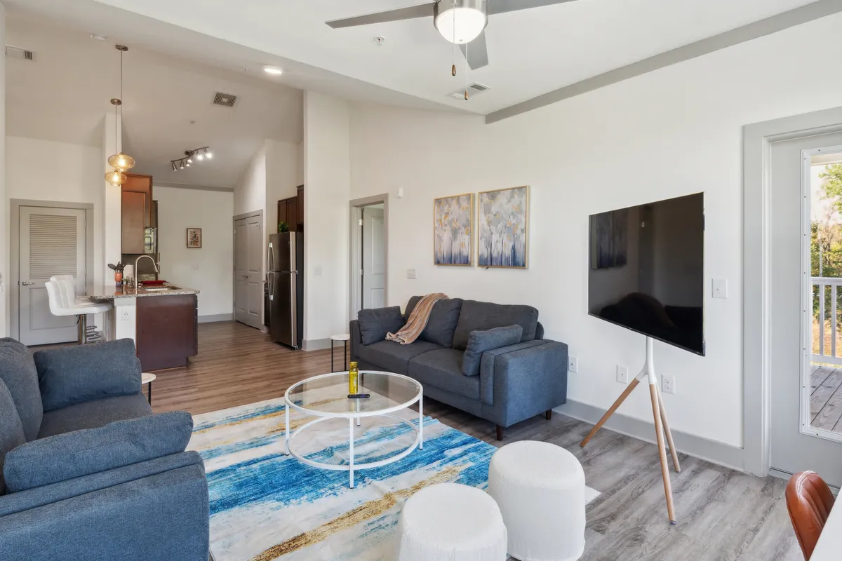 This image features a cozy and inviting living area with a sofa, coffee table, artwork on the wall, a television, and an open kitchen area visible in the background. The space is styled with modern décor and soft lighting, creating a welcoming atmosphere ideal for relaxation and entertaining.