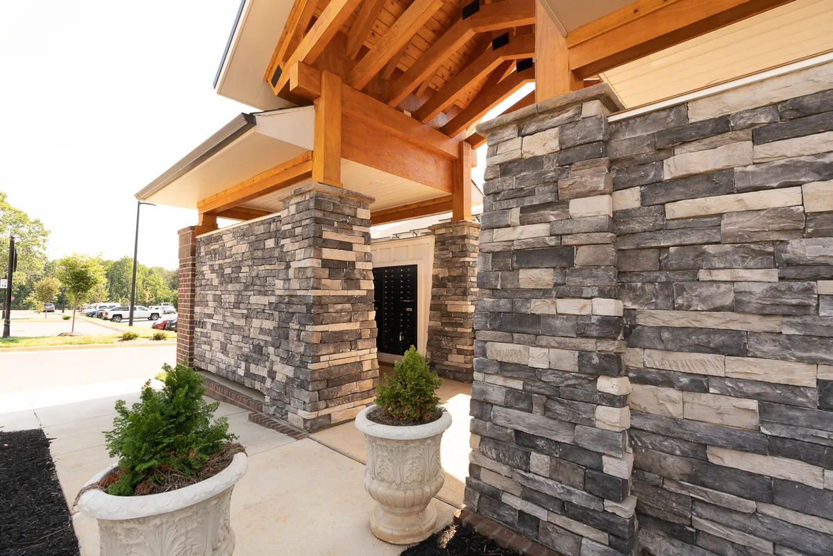 A close-up of the inviting stone walls and wooden beams at the building’s entrance, accented by planters. The scene is warm and inviting, emphasizing moments of welcome and arrival.