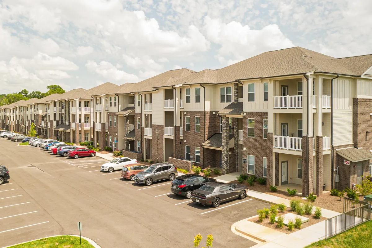 Rows of apartment buildings framed by landscaped greenery and parked cars, suggesting convenient parking and vibrant communal spaces for residents. The environment feels active, safe, and community-focused.