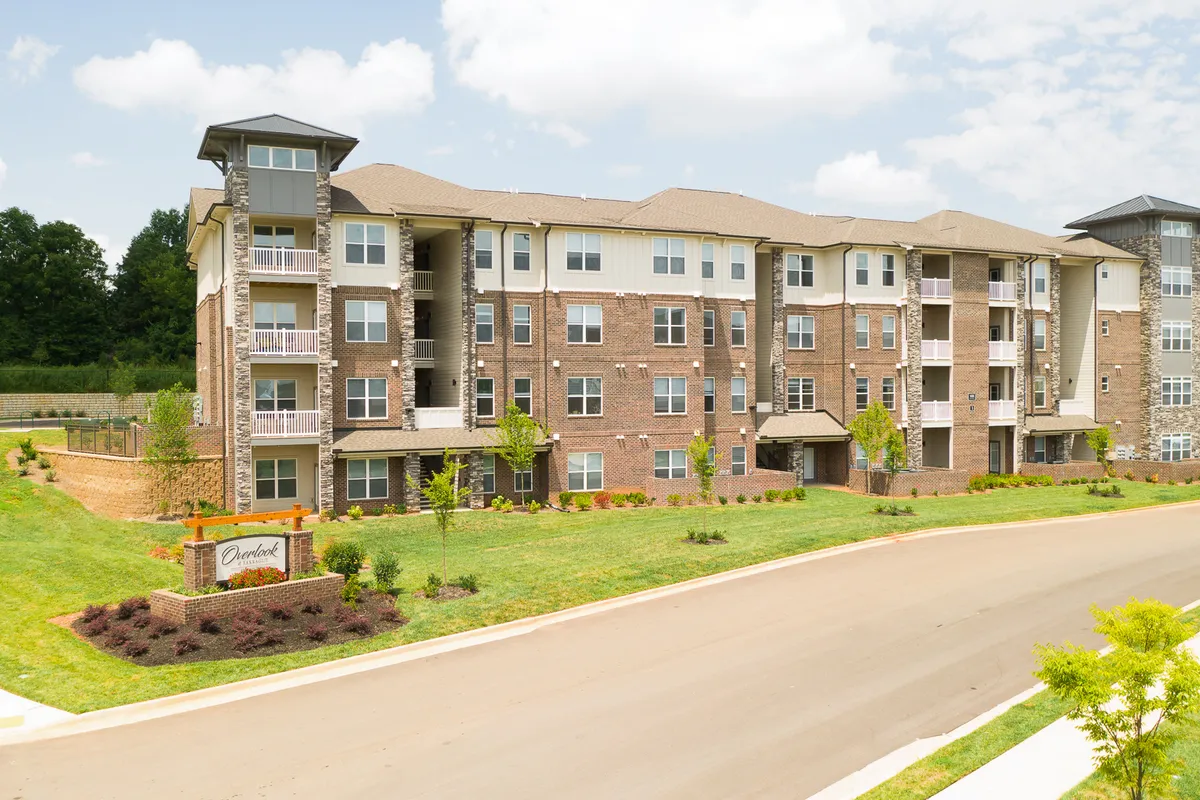 A modern apartment building surrounded by lush green landscaping, with clear signage and open communal garden spaces. Residents can be seen relaxing outdoors, enjoying the sunshine and the inviting atmosphere of the property.