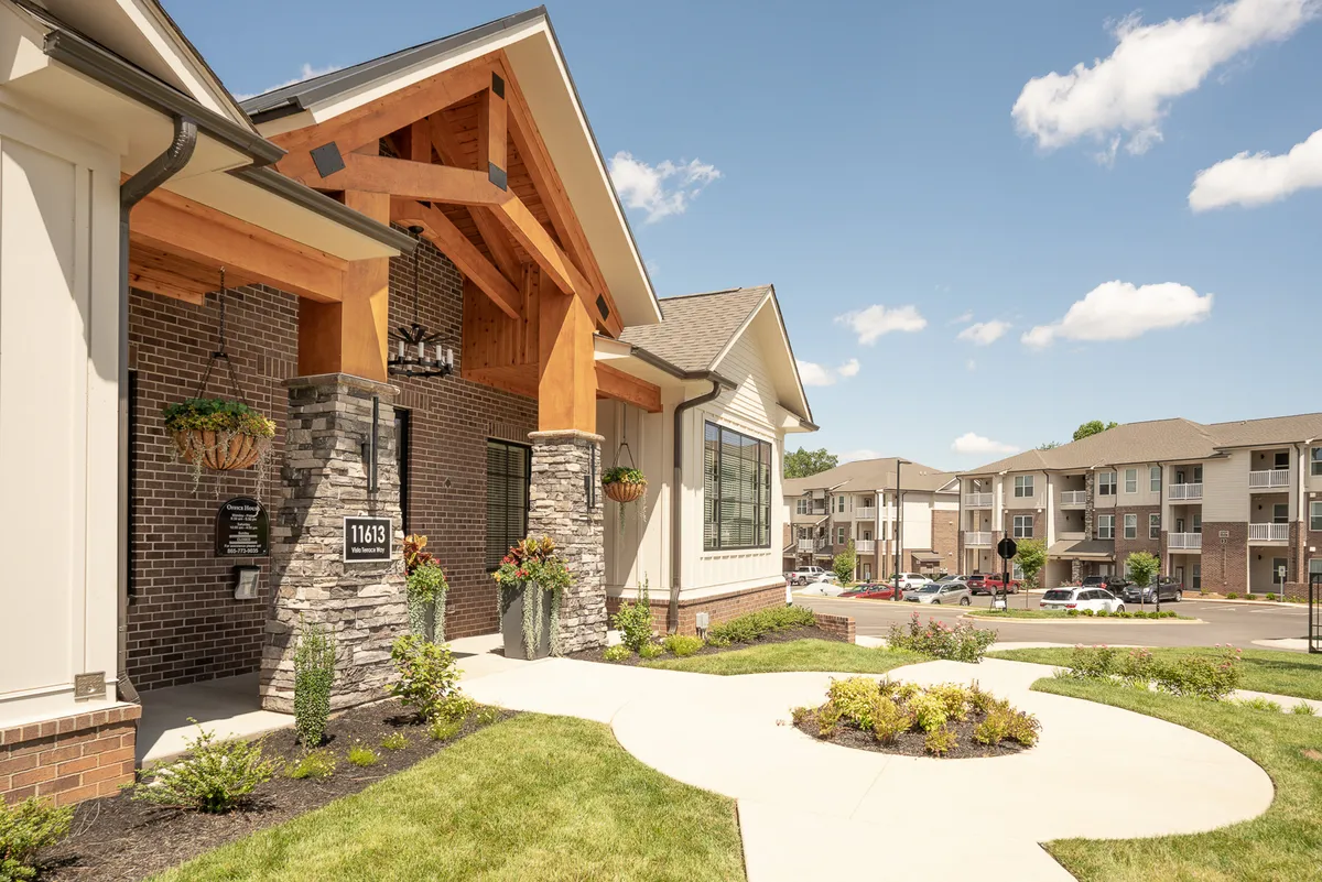 This image shows the exterior of the property with a striking wooden entryway, a large overhang, and a stone facade surrounded by lush landscaping. Large windows suggest bright, inviting interiors that blend seamlessly with the welcoming outdoor environment.